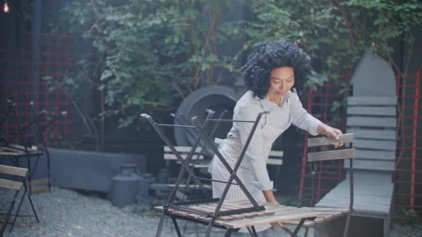 午前中にテラスで働くアフリカ系アメリカ人のウェイトレス 仕事を始める前に良い感情を持っているカーリーヘアの忙しい女性 ストリートカフェの床下の椅子の配置 — ストック動画