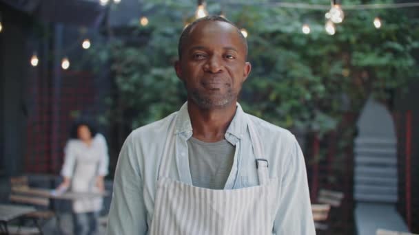 カメラの前に立っている間エプロンを身に着けているアフリカ系アメリカ人の男性ビジネスオーナーの肖像画 ハンサムな男が手を横切って ポジティブに微笑んでいる 個人のカフェを開店することを誇りに思います — ストック動画