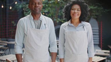 Kameranın önünde dururken önlük takan Afrikalı Amerikalı işçilerin portresi. Tatmin olmuş garsonlar el ele tutuşurken birbirlerine bakıyorlar. Kafede birlikte çalışıyoruz. Mutlu insanlar kavramı.