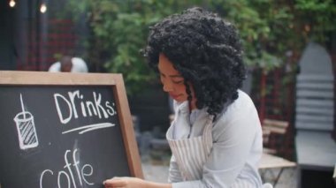 Güzel Afro-Amerikalı kadın, insanların restorana gelmesi için kömürle yazı yazıyor. Kafenin neler sunabileceğini görmek için farklı türde içki yazan bir kız..