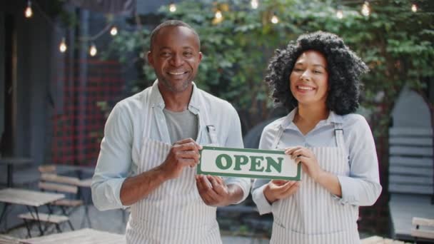 2人のアフリカ系アメリカ人ウェイターとエプロンがカフェテラスでオープンサインを保持しています 幸せな労働者はバーの前に立っている間喜んで微笑んでいます 顧客を受け取る準備をする 労働日のコンセプト — ストック動画