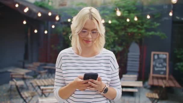 カメラの前に立っている間 彼女の携帯電話を使用している若い白人女性 スマートフォンのメッセージを誰かにタイピングします ソーシャルメディアで友達とチャットしながら陽気な笑顔 リモートコミュニケーション — ストック動画