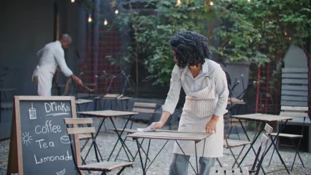 勤勉なアフリカ系アメリカ人は 仕事を始める前にテーブルを拭く 朝のテラスを掃除するために同僚を助ける忙しい女性 若いスタッフがカフェを掃除しています パートナーシップコンセプト — ストック動画
