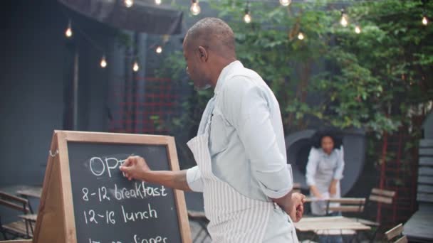 アフリカ系アメリカ人男性がメッセージを発信している フレンドリーな男性オーナーは カフェの食べ物を試すために新しい顧客を歓迎します ぼやけた背景で目に見えるハードワークウェイトレスクリーニングテラスゾーン — ストック動画