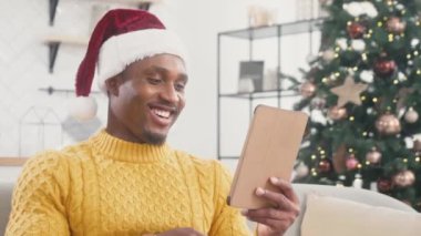 Odadaki kanepede tablet cihazıyla dinlenirken Noel Baba şapkası takan tatmin olmuş Afrikalı Amerikalı adam. Teknoloji aletinde komik videolar izliyor. Noel 'de eğleniyoruz. Tatil kavramı.