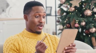 Sarı örgü süveter giyen genç, çekici Afrikalı Amerikalı adam Noel için tabletinden alışveriş yapıyor. Tatmin olmuş erkek kredi kartı tutuyor ve ödemeden dolayı mutlu bir şekilde gülümsüyor..