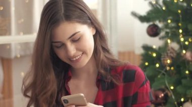 Dişli gömlekli güzel beyaz kız cep telefonuyla internetten iletişim kuruyor. Noel ağacının yanında çelenk ile otururken arkadaşlarına telefonda mesaj yazıyormuş. Teknoloji kullanılıyor.