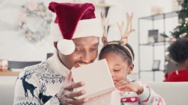 Yakışıklı Afrikalı Amerikalı baba koltuğunda çocuğunun kucağında oturuyor. Geyik boynuzu halkalı küçük çocuk ışık saçan bir kutu açıyor. Mutlu çocuk Noel süslemesini hediyeyle çıkarıyor..