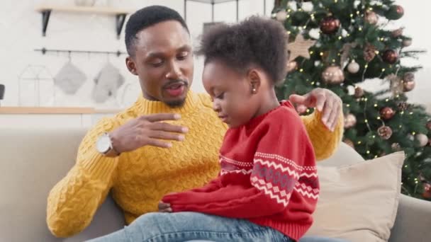 魅力的なアフリカ系アメリカ人の父親は バックグラウンドでクリスマスツリーで居心地の良い部屋でソファーに従順な娘と休んでいます 父親は彼の膝の上に座っている間 子供と話した ファミリーデイコンセプト — ストック動画