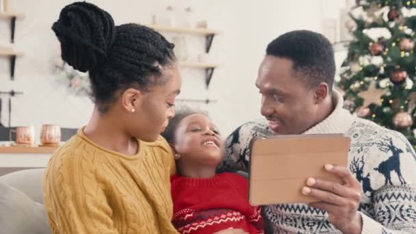 満足しているアフリカ系アメリカ人の両親は 子供が自宅の現代のリビングルームでタブレットデバイスでビデオ通話をしている バックグラウンドでクリスマスツリーでカメラで話して笑顔で幸せな家族 — ストック動画