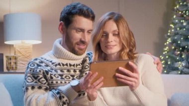 Arka planda Noel ağacı ile oturma odasındaki kanepede dinlenirken kalın örülü süveter giyen beyaz çifte bayılıyorlar. Evde yaygın dinlenme sırasında tablet aygıt kullanan pozitif genç aile