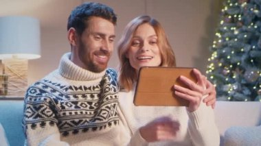 Beyaz bir çiftin Noel ağacıyla oturma odasındaki kanepede otururken tablet cihazında görüntülü konuşma yapması. Noel arifesinde mutlu aile iletişimi. Uzak neşeli sohbet.