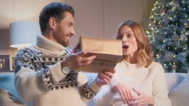 Yakışıklı beyaz koca, sevgili karısına Noel ağacından önce koltukta otururken sürpriz yapıyor. Tatilde hediye alan hoş bir kadın. Mutlu bir aile yeni yılı ya da Noel 'i birlikte kutluyor.