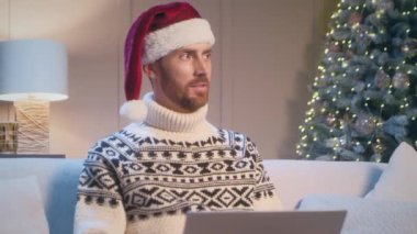 Noel Baba şapkası ve sıcak süveter giyen beyaz bir adam Noel ağacının yanındaki kanepede çelenkle otururken. Tebrik mesajı yazan genç adam. Noel tatili ya da Yeni Yıl.