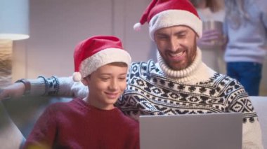 Oturma odasında kanepede otururken oğlu Noel Baba şapkası takan yakışıklı beyaz bir baba. Mutlu aile videoları ya da dizüstü bilgisayarda komik filmler izliyor. Noel arifesinde boş vakit geçirmek..