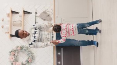 Dikey videoda Afrikalı Amerikalı bir babanın küçük tatlı kızıyla modern beyaz mutfakta geyik boynuzu çemberinde dans ettiğini görüyoruz. Mutlu bir aile Noel örgü süveteri giyer ve dinlenir..