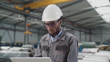 Güvenlik kaskı ve üniforması olan sakallı Afro-Amerikan adam büyük bir atölyede dizüstü bilgisayarında çalışıyor. Genç mühendis dijital cihazında endüstriyel fabrikada yeni üretim hattı tasarladı..