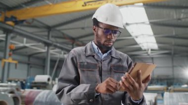 Çekici Afrikalı Amerikalı bir adam, büyük bir endüstriyel atölyede beklerken müşteriyle tablet cihazında uzaktan sohbet ediyor. Fabrikada çalışırken dijital aygıt kullanmak.