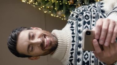 Noel ağacının yanında duran, kalın kazak giyen yakışıklı beyaz adamın dikey oryantasyonu. Çevrimiçi iletişim kurarken veya sosyal medyada gezinirken akıllı telefon kullanan sakallı erkek.