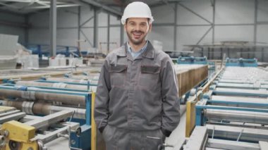 Fabrikadaki endüstriyel makinelerin arasında duran üniformalı tatmin olmuş profesyonel bir adama zum yapıyorum. Mutlu işçi ellerini cebinde tutarken kameraya olumlu gülümsüyor. İşyeri.