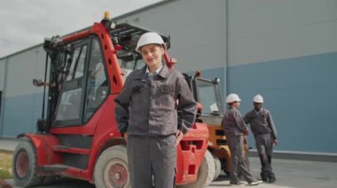 İş üniforması giyen güzel bir kız ellerini cebine sokuyor. Arka planda, forklift yanında dikilirken konuşan iki melez adam var. İşçiler fabrikada malları boşaltmak için bekliyorlar..