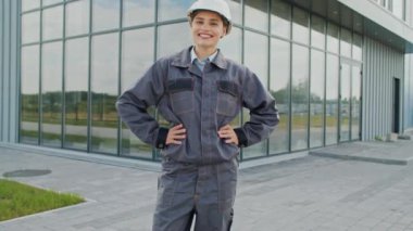 Beyaz kasklı güzel beyaz kadın kameraya bakarken neşeyle gülümsüyor. Modern cam bina önünde dururken mutlu kadın mühendisin iyi duyguları var. Dışarıda çalışıyorum..