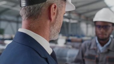 Fabrikada çalışırken güvenlik ekipmanı takan melez insanların yan görüntüsü. Çalışkan ustabaşı yönetmeniyle konuşuyor. Adamlar depoda dikilirken iletişim kuruyor..