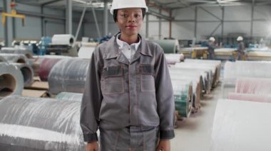 Gri üniformalı ve kasklı Afro-Amerikan kadın işçi depoda metal eşyaların yanında duruyor. Modern fabrikada kameraya gülümseyen genç bir kadın. Üretim süreci.