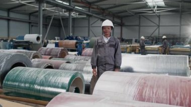 Üniformalı ve güvenlik ekipmanlı mutlu Afro-Amerikan kadın imalat ürünleri arasında duruyor. Üretimin kontrol hacmi. Arka planda iki adam mal üretimi sırasında konuşuyor..