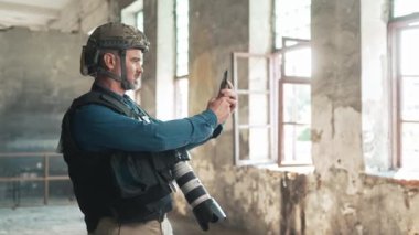 Gri sakallı beyaz bir erkeğin haberlere ya da işle ilgili bilgilere ulaşmak için mobil cihazını kullandığına yakından bakın. Askeri çatışma bölgesinde çalışıyor. Terk edilmiş binada saklanan basın çalışanı.