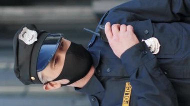 Dikey çekim. Açık havada. Koruyucu maskeli yakışıklı polisin portresi sokakta kalırken ciddi ve kendinden emin görünüyor. Tıbbi maskeli bir polis kameraya bakıyor. Sosyal güvenlik kavramı.