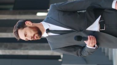 Modern şirket binasının önünde duran iyi giyimli ofis yöneticisinin dikey çekimi. Yakışıklı gazeteci gülümsüyor ve modern ticari tesisin dışındaki kameraya bakıyor..