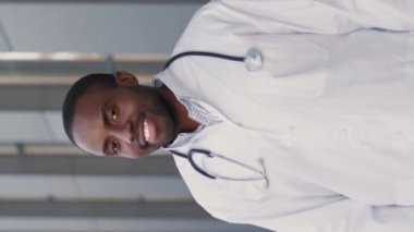 Merkez hastanesi yakınlarındaki şehir sokağında duran koruyucu elbise giyen ve poz verirken kameraya gülümseyen neşeli çok ırklı doktorun dikey çekimi. Genç profesyonel tıp çalışanı..