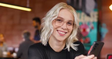 Kafedeki masada otururken akıllı telefonunu tutan, daha iyi görebilmek için gözlüklü beyaz kadın portresi. Güzel kız barda dinlenirken iyi hissediyor. Neşeli gülümseme kameraya.