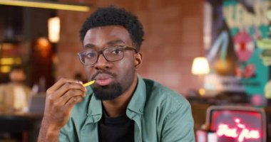 Kafede ya da barda otururken gözlüklü Afro-Amerikan bir adamın portresi. Genç adam fast food seviyor. Evde yemek yerken kameraya ciddi ciddi bakıyorum. Beslenme kavramı.