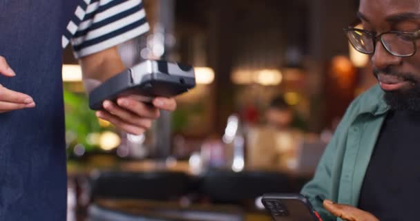 支払い端末を保持しているウェイトレスのカメラビュー アフリカ系アメリカ人の男性が電話で支払っている カフェで食べ物や飲み物を買うために携帯電話を使っている若者 テクノロジーデバイスの購入 — ストック動画