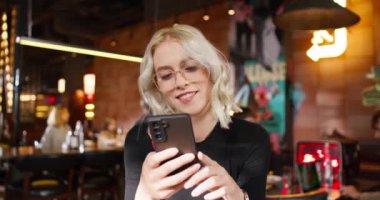 Gözlüklü genç beyaz kadın telefonunu tutarken etrafına bakıyor. Güzel bir kadın kafede otururken akıllı telefondan biriyle uzaktan iletişim kuruyor. Teknoloji kavramı.