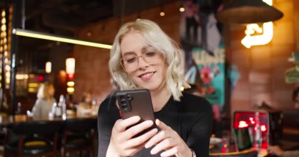 携帯を持っている間 眼鏡をかけた若い白人女性 カフェに座っている間 スマートフォンで誰かと遠隔でコミュニケーションする美しい女性 テクノロジーの概念 — ストック動画