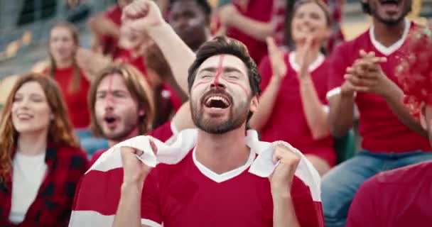 积极的球迷们为他们的球队欢呼喝彩 英俊的男子举着英国国旗 对着镜头微笑 观众为胜利感到高兴 球迷们在体育场玩得很开心 体育的概念 — 图库视频影像