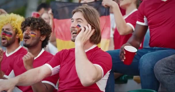 長い髪と青い目を持つひげ付きの白人男性のカメラビュー スタジアムトリプルーンに座ってドイツ代表をサポートしながら 彼のモバイルデバイスで感情的に話す男 — ストック動画
