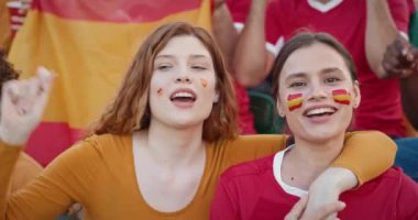 Yanaklarında İspanyol ulusal bayrakları olan birkaç beyaz kadın. Kızlar en sevdikleri takımı desteklemek için stadyum tribünlerinde birbirlerine sarılıp şarkı söylüyorlar..