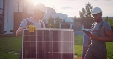 İki çok kültürlü adam yenilenebilir enerji kuruluşunda çalışıyor. Çimlerde bekleyen çok ırklı müteahhitler. Erkek teknisyenler güneş panelleri kuruyor. Afro-Amerikalı adam elinde tablet tutuyor..