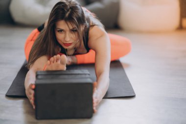 Genç, güzel, kızıl saçlı bir atlet spor yapıyor, evde bloklu bir spor minderi üzerinde esneme egzersizleri yapıyor. O, turuncu bir eşofman giyiyor. Yakın plan..