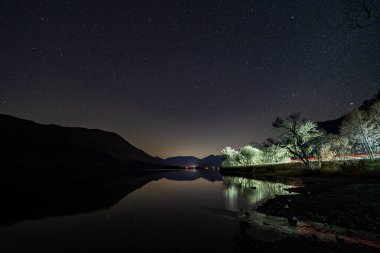 İngiliz Göl Bölgesi 'ndeki Ullswater' ın üzerindeki gece gökyüzü, geçen bir araba tarafından aydınlatılmış ağaçlar.
