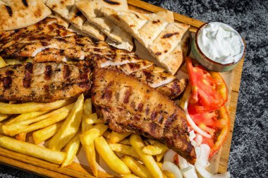 Mükemmel ızgara tavuk göğsü ve tuzlu domuz dilimleri, yanında da altın patates kızartması, taze domates ve yoğurt sosu. Hepsi ustalıkla sade bir tahta üzerine yerleştirilmiş..