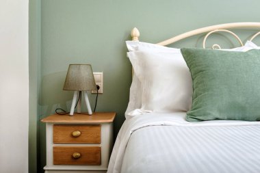 Beyaz ve zeytin tonlarının renk şemasını içeren çağdaş bir yatak odası konsepti. Görüntü, yatağın bir kısmını, üç ayaklı lambayla süslenmiş ahşap bir komodinin yanında sergiliyor.