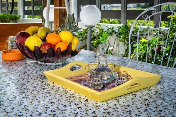 stock image Set summer table featuring vase filled with a variety of fresh fruits including apples, bananas, oranges, lemons. Tray with carafe filled with clear white spirit, raki, accompanied by small glasses