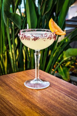 Açık sarı bir kokteyl ile doldurulmuş Coupe bardağı kenarında taze turuncu bir kesikle güzelce süslenmiş. Kokteyl klasik bir coupe bardakta servis edilir, canlı rengini vurgular..