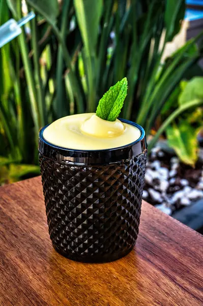 stock image Thickened black glass cup filled with cocktail, topped with layer of yellow foam, garnished with fresh mint leaf. Dark, opaque glass contrasts with vibrant yellow foam, creating striking visual.