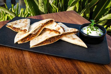 Meksika 'dan esinlenilmiş, mükemmel düzenlenmiş altın-kahverengi quesadilla dilimli siyah tabakla yakın plan bir fotoğraf. Tabağın yanında, küçük siyah kase kremalı beyaz sosu barındırır. 